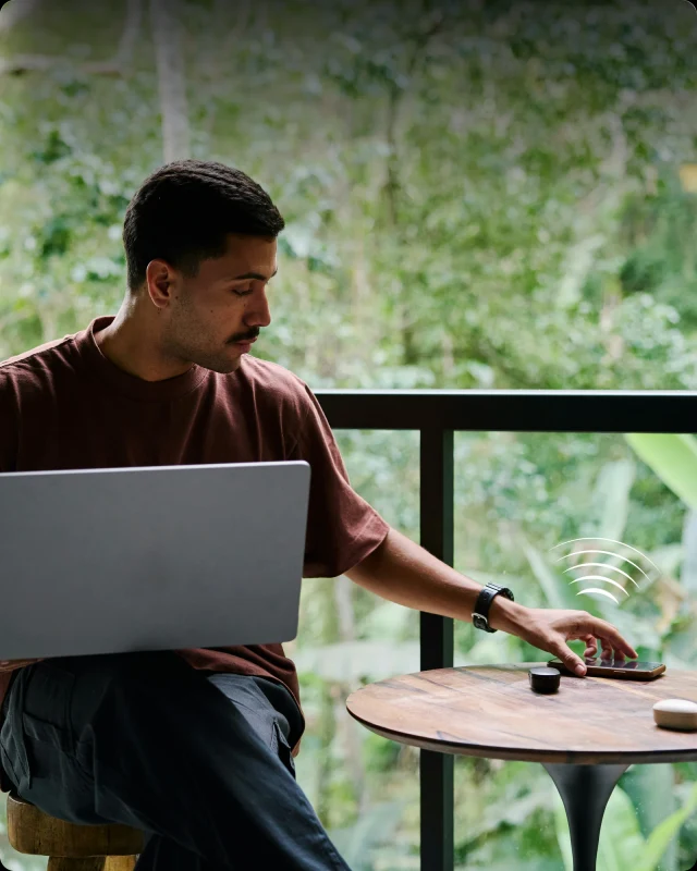 Een man op een balkon met een laptop op zijn schoot, die zijn deur ontgrendelt met de telefoon op tafel